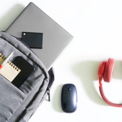 Gray urban backpack or bag with laptop, external ssd drive, mobile phone, notepad and pen on a white table next to mouse and red headphones. Tools and gadgets of a student or freelancer. Close-up